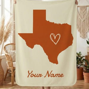 Peut inclure: Couverture crème avec un contour orange brûlé de l'État du Texas et un cœur blanc au centre. Les mots "Your Name" sont écrits en cursive en bas. La couverture est tenue par une personne.