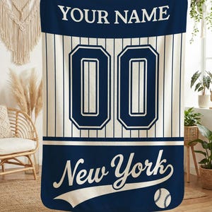 May include: A navy blue and cream-colored baseball-themed blanket. The blanket features the text "YOUR NAME" at the top, the number "00" in the center, and "New York" with a baseball graphic at the bottom. Vertical stripes run down the center.