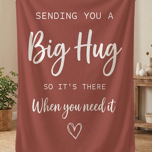 Könnte beinhalten: Eine rostfarbene Decke mit weißer Schrift, die lautet: "SENDING YOU A Big Hug SO IT'S THERE When you need it" und ein Herz. Die Decke wird von einer Person gehalten.