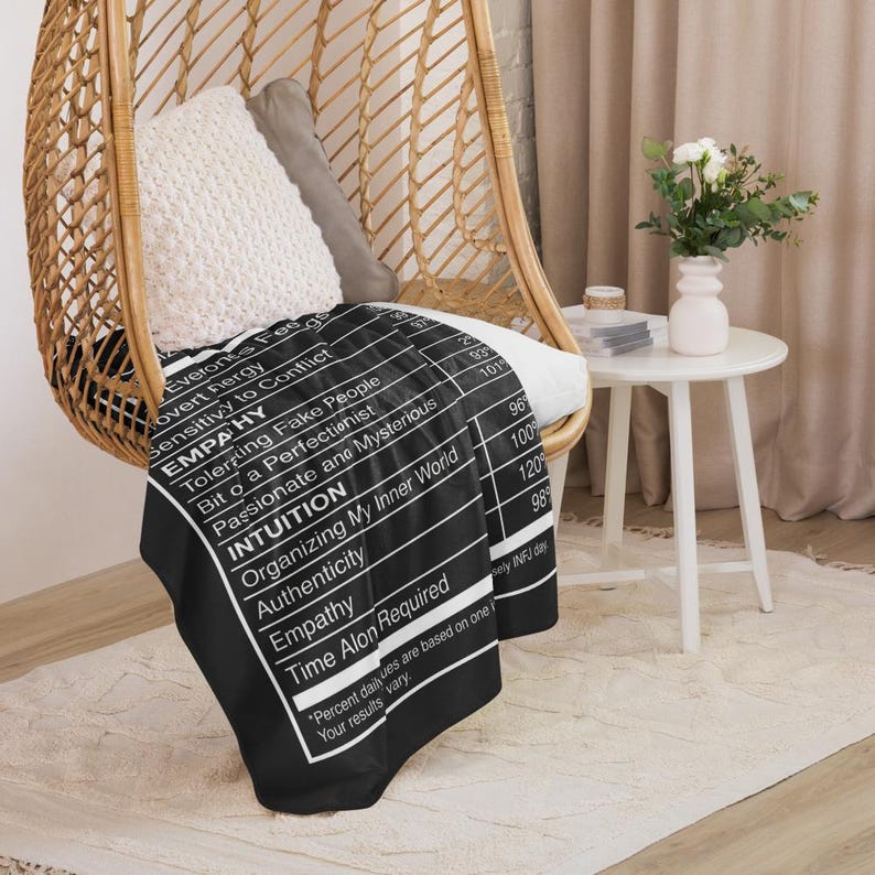 May include: A black throw blanket with white text, draped over a chair. The text lists traits like "Empathy" and "Intuition" in a nutrition facts style. The blanket is on a woven chair with white pillows.