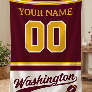 May include: Maroon and gold personalised blanket featuring the text "YOUR NAME" above two gold numbers. Below, the word "Washington" is displayed in a script font, along with a football graphic. The blanket has a white border.