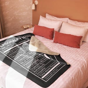 May include: A black blanket with white text listing "Personality Facts" and traits like "Empathy" and "Intuition." The blanket is on a bed with white and red pillows. A beige throw is draped over the blanket.