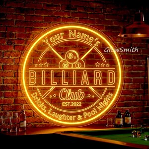 Può includere: Un'insegna al neon per un biliardo, che brilla con una calda luce gialla contro un muro di mattoni. L'insegna circolare presenta il testo "Your Name's BILLIARD Club EST.2022" con palle e stecche da biliardo. Sono incluse anche le parole "Drinks, Laughter & Pool Nights".