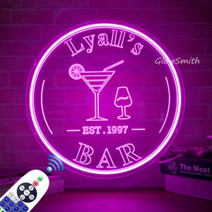May include: A circular neon sign glowing in bright pink, featuring the text Lyall's BAR and EST. 1997. The design includes a martini glass with a citrus slice and a small wine glass. A remote control with various buttons is visible in the foreground.