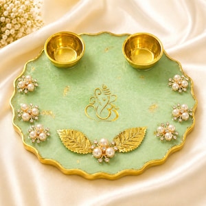 Op de afbeelding: Een decoratief groen dienblad met gouden rand, met twee kleine gouden kommen. Het dienblad is versierd met gouden bladaccenten, bloemversieringen van parels en strass, en een gouden Ganesh-symbool. Ontworpen voor religieuze of decoratieve doeleinden.