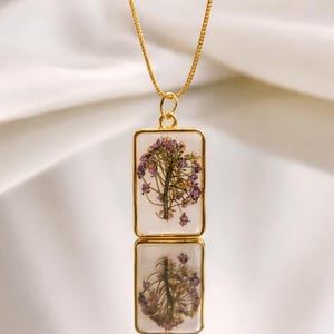 Könnte beinhalten: Goldfarbene Halskette mit rechteckigem Anhänger. Der Anhänger enthält ein Arrangement aus getrockneten lila und weißen Blumen, das in klarem Harz eingeschlossen ist. Die Halskette wird auf einer reflektierenden Oberfläche präsentiert.