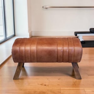 May include: A brown leather bench with a cylindrical top and wooden legs. The bench features vertical stitching and a warm colour. The angled wooden legs provide stability. The bench is likely used for seating or as a decorative item.