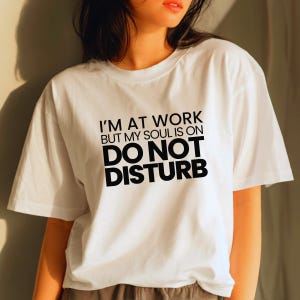 May include: White t-shirt with the text "I'M AT WORK BUT MY SOUL IS ON DO NOT DISTURB" in black. The shirt has short sleeves and a crew neck. The text is centered on the front of the shirt.