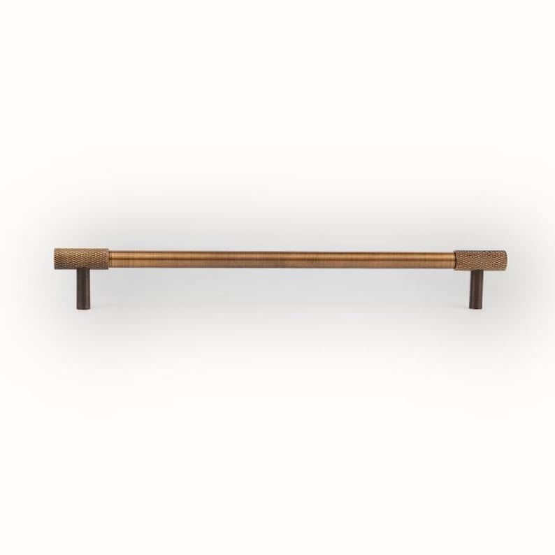 Puede incluir: Un tirador de armario de color bronce con extremos texturizados. El tirador es una barra larga y recta con un centro cil&iacute;ndrico y extremos rectangulares. Los postes de montaje son de color marr&oacute;n oscuro. Aproximadamente 20 cm de largo.