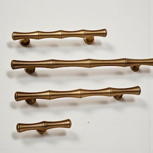 Puede incluir: Cuatro tiradores de armario de color latón antiguo en varios tamaños. Los tiradores tienen un diseño de estilo bambú, con una serie de segmentos cilíndricos y extremos redondeados. Están montados sobre una superficie blanca.