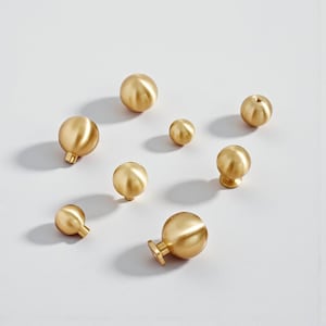 May include: A collection of brass-colored cabinet knobs in various sizes and designs. The knobs are spherical, with some featuring a stem or a decorative base. The hardware is arranged on a white surface, showcasing their metallic finish.