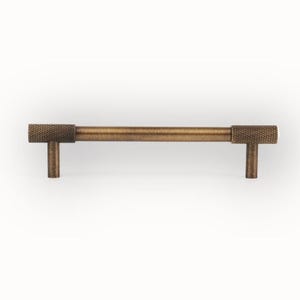 Puede incluir: Un tirador de armario de color bronce con un mango cil&iacute;ndrico texturizado y dos extremos en forma de T. El herraje tiene una est&eacute;tica vintage. El tirador se muestra sobre un fondo blanco. Mide aproximadamente 15 cm de largo.