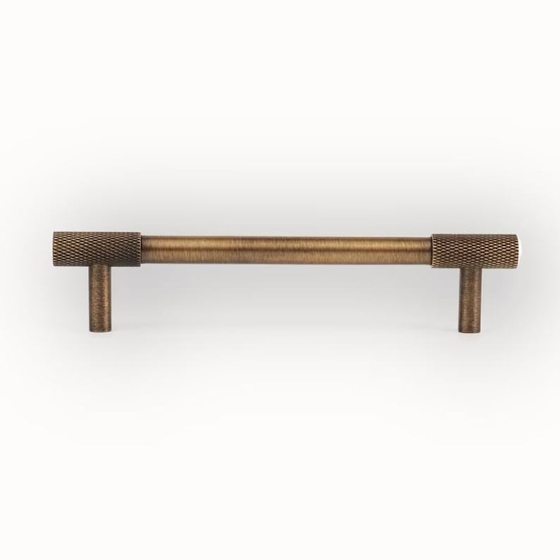 Puede incluir: Un tirador de armario de color bronce con un mango cil&iacute;ndrico texturizado y dos extremos en forma de T. El herraje tiene una est&eacute;tica vintage. El tirador se muestra sobre un fondo blanco. Mide aproximadamente 15 cm de largo.