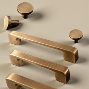 Może przedstawiać: Zestaw mosiężnych okuć do szafek, w tym kilka prostokątnych uchwytów i okrągłych gałek. Okucia mają szczotkowane wykończenie i nowoczesny design, odpowiednie do szafek kuchennych lub łazienkowych.