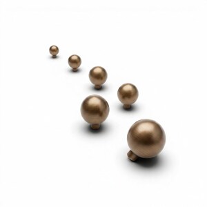May include: Six bronze-colored spherical cabinet knobs arranged diagonally on a white background. Each knob has a small, cylindrical base. The knobs vary slightly in size, with the largest one in the foreground.
