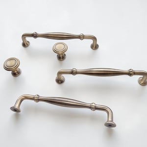 May include: A collection of five cabinet hardware pieces in an antique brass finish, consisting of three elongated arched drawer pulls with rounded ends and two matching circular knobs, all featuring a smooth, metallic, brushed texture.