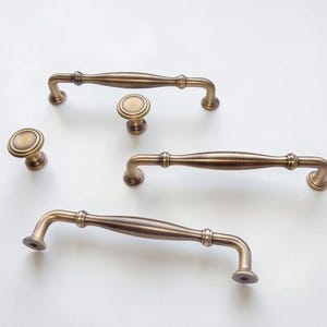 May include: A collection of antique brass hardware consisting of two round cabinet knobs and three arched drawer pulls. Each piece features a smooth, metallic finish with a subtle brushed texture and rounded, tiered detailing at the mounting points.
