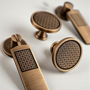 May include: Close-up of antique bronze cabinet hardware. The set includes two oval knobs, a round knob, and a rectangular handle. Each piece features a textured, geometric pattern and a warm, metallic finish.