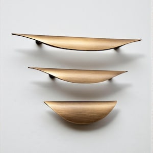 May include: Three brass-coloured cabinet pulls of different sizes are displayed against a white backdrop. Each pull has a curved, leaf-like form with a slight sheen. The hardware is designed for a modern aesthetic, suitable for various furniture pieces.