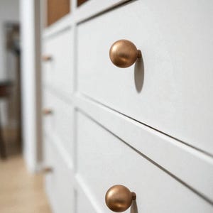 May include: Close-up of a white cabinet with several drawers. Each drawer has a round, bronze-colored knob. The knobs are evenly spaced and create a symmetrical pattern. The cabinet is in focus, with a blurred background.