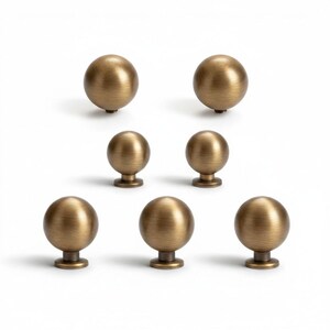 May include: Seven antique brass-colored cabinet knobs arranged on a white background. The knobs are spherical with a short, cylindrical base. The finish has a slightly aged appearance, suggesting a vintage or classic style. Ideal for furniture restoration or adding a touch of elegance.