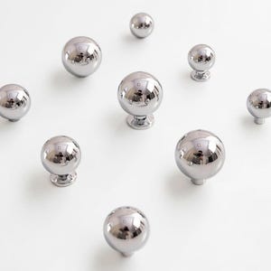 May include: A collection of shiny, chrome-finished cabinet knobs. The knobs are spherical, reflecting the surrounding environment. Some knobs have a small base, while others are simple spheres. The knobs are arranged on a white surface.