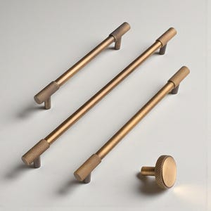 May include: Four bronze-colored cabinet hardware pieces. Three are long bar pulls with textured ends, and one is a round knob. The hardware has a warm, metallic finish, suitable for kitchen or bathroom cabinets.