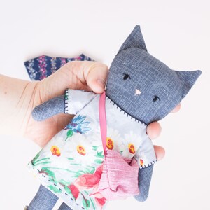 Peut inclure: Poup&eacute;e chat grise faite &agrave; la main, v&ecirc;tue d'une robe &agrave; fleurs, d'un sac &agrave; main rose et de chaussures dor&eacute;es. La poup&eacute;e a des yeux cousus noirs et un nez rose. La robe pr&eacute;sente un motif de marguerites blanches et de fleurs rouges.