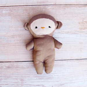 May include: A brown plush monkey toy with a white face and embroidered eyes and nose. The monkey is standing on a light wood background.