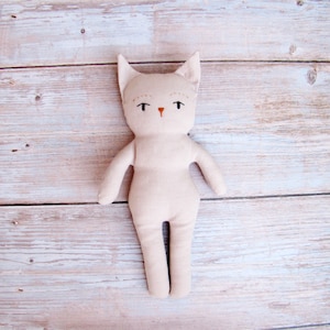 Peut inclure: Poup&eacute;e chat beige faite &agrave; la main, avec des traits de visage brod&eacute;s, dont des yeux noirs et un petit nez orange. La poup&eacute;e a un design simple et minimaliste avec un corps en tissu doux et des bras tendus. La poup&eacute;e est pos&eacute;e sur une surface en bois clair.