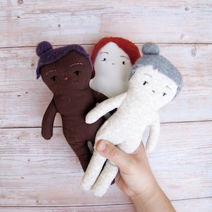 May include: Three handmade fabric dolls with embroidered faces. The dolls are brown, white, and gray with different colored hair. The dolls are sitting on a light wood surface.