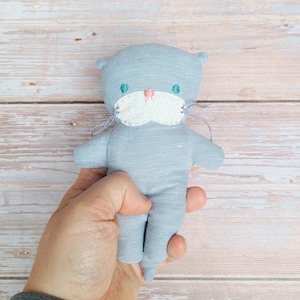 Könnte beinhalten: Eine handgefertigte blaue Katzendoll mit gestickten Details. Die Puppe hat ein weißes, gesticktes Gesicht mit rosa und grünen Akzenten und lange graue Schnurrhaare. Die Puppe ist aus weichem Stoff gefertigt und hat kleine Arme und Beine.