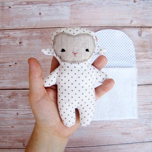 Puede incluir: Un peluche de cordero blanco y gris con lunares, con una cara de fieltro marrón y ojos negros. El cordero está siendo sostenido en una mano y está sentado sobre un fondo de tela blanca.