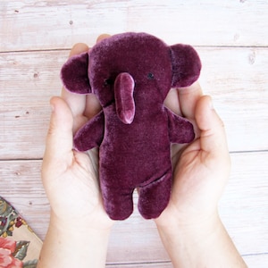 May include: A plush purple elephant toy with a long trunk and small black eyes. The toy is being held in the hands of a person.