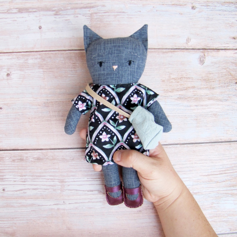 Peut inclure: Poup&eacute;e chat grise faite &agrave; la main, v&ecirc;tue d'une robe &agrave; motifs avec des fleurs roses et d'un petit sac bleu. La poup&eacute;e a des chaussures violettes et une sangle beige. La poup&eacute;e chat est tenue dans la main d'une personne.