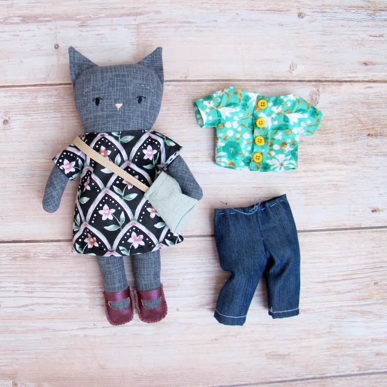 Peut inclure: Une poup&eacute;e chat grise v&ecirc;tue d'une robe noire &agrave; motifs floraux, de leggings gris et de chaussures bordeaux. La poup&eacute;e a un sac assorti, une chemise &agrave; fleurs verte s&eacute;par&eacute;e et un jean bleu.