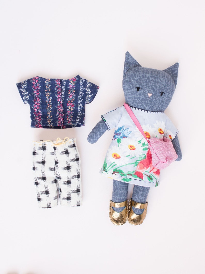 Peut inclure: Une poup&eacute;e chat bleue faite &agrave; la main avec une robe &agrave; fleurs, un sac &agrave; main rose et des chaussures dor&eacute;es. La poup&eacute;e a un haut &agrave; fleurs assorti et un short &agrave; carreaux noir et blanc. La tenue de la poup&eacute;e est con&ccedil;ue pour le jeu et l'imagination.