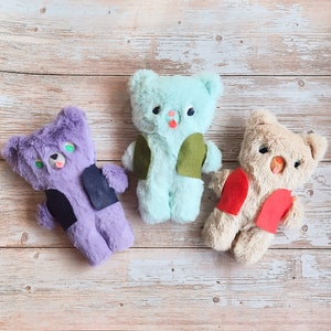 Puede incluir: Tres osos de peluche con pelaje de diferentes colores. El primer oso es azul claro con un chaleco verde, el segundo oso es morado con un chaleco negro y el tercer oso es marrón claro con un chaleco rojo.