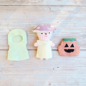 May include: Three handmade felt Halloween decorations: a light green ghost costume, a mushroom person with a pink and white spotted cap, and an orange pumpkin with a black jack-o'-lantern face. The items are arranged on a light wood grain surface.