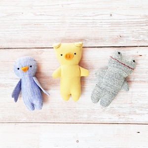 Puede incluir: Tres juguetes de peluche hechos a mano: un cerdo amarillo, un pájaro azul y una rana gris. Los juguetes están sobre una superficie de madera blanca.