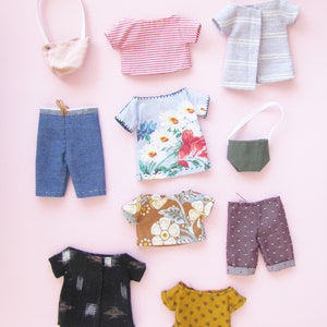 Peut inclure: Une collection de v&ecirc;tements de poup&eacute;e miniatures, comprenant des hauts, des pantalons et un sac &agrave; main, dispos&eacute;s sur un fond rose. Les v&ecirc;tements pr&eacute;sentent divers motifs et couleurs, tels que des rayures, des imprim&eacute;s floraux et des pois. L'ensemble comprend un short en jean et un petit sac &agrave; main rose.