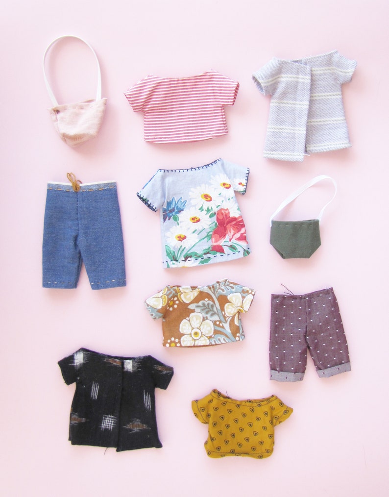 Peut inclure: Une collection de v&ecirc;tements de poup&eacute;e miniatures, comprenant des hauts, des pantalons et un sac &agrave; main, dispos&eacute;s sur un fond rose. Les v&ecirc;tements pr&eacute;sentent divers motifs et couleurs, tels que des rayures, des imprim&eacute;s floraux et des pois. L'ensemble comprend un short en jean et un petit sac &agrave; main rose.