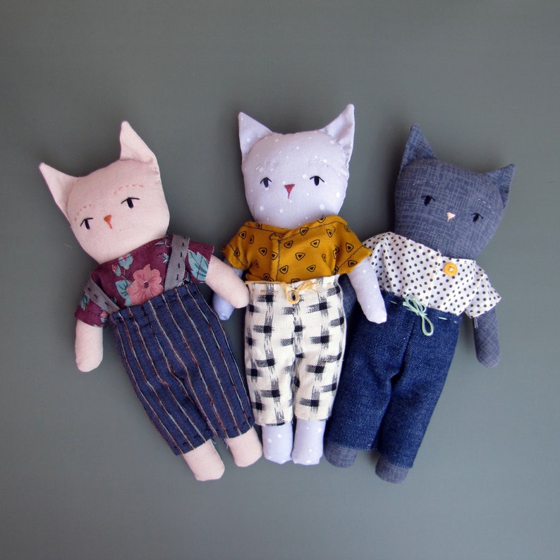 Peut inclure: Trois poup&eacute;es chats faites &agrave; la main avec des v&ecirc;tements en tissu. Les poup&eacute;es ont des oreilles de chat, des visages brod&eacute;s et sont habill&eacute;es de diff&eacute;rentes tenues, dont des salopettes, des chemises &agrave; motifs et des pantalons. Les poup&eacute;es sont dispos&eacute;es sur un fond vert uni.