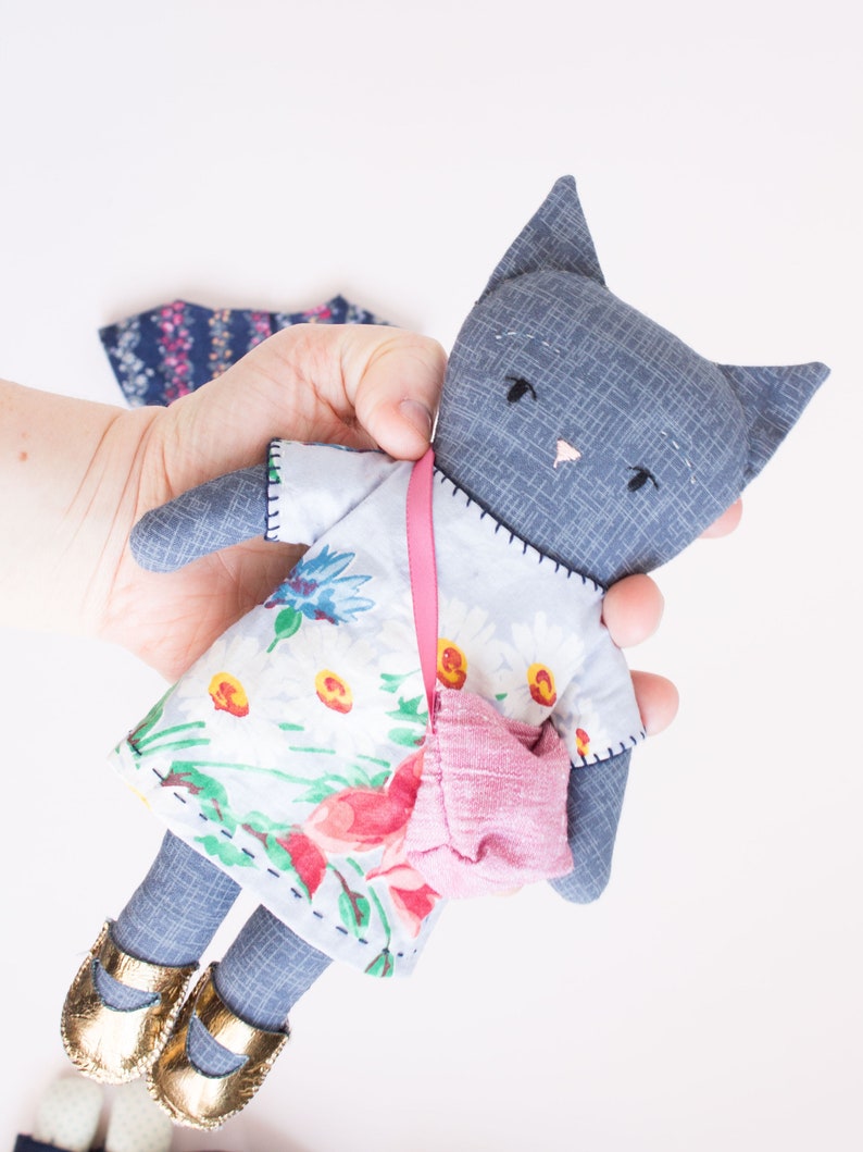 Peut inclure: Poup&eacute;e chat grise faite &agrave; la main, v&ecirc;tue d'une robe &agrave; fleurs, d'un sac &agrave; main rose et de chaussures dor&eacute;es. La poup&eacute;e a des yeux cousus noirs et un nez rose. La robe pr&eacute;sente un motif de marguerites blanches et de fleurs rouges.