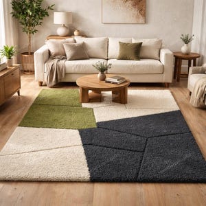Puede incluir: Una alfombra geométrica con un diseño moderno. La alfombra presenta secciones verdes, blanquecinas y gris oscuro. Está colocada sobre un suelo de madera en un salón, con un sofá blanco y una mesa de centro de madera.