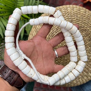 Puede incluir: Un collar de cuentas blancas rectangulares ensartadas en un cordón blanco. Las cuentas tienen sutiles variaciones de color y textura. El collar se muestra sobre un fondo neutro, resaltando los tonos naturales de las cuentas.