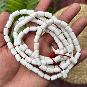 Puede incluir: Un collar de cuentas blancas rectangulares con bordes redondeados, ensartadas en un cordón rojo. Las cuentas se sostienen en una mano, con un fondo neutro. Las cuentas parecen estar hechas de un material natural.