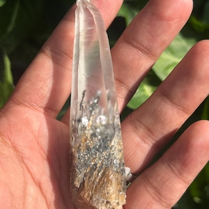 May include: A clear, elongated quartz crystal point with a pointed tip. The base of the crystal features a dense, textured inclusion of dark grey, black, and golden-brown mineral deposits that transition into a transparent, colourless upper section.