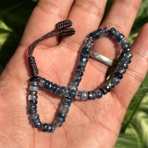 Puede incluir: Una pulsera compuesta por pequeñas cuentas facetadas y translúcidas de color azul y gris dispuestas en círculo. Las cuentas están ensartadas en un cordón marrón oscuro con un cierre deslizante ajustable y dos extremos pequeños anudados.