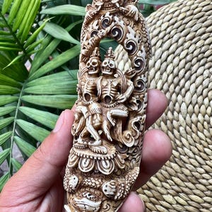 Könnte beinhalten: Eine detaillierte, cremefarbene Schnitzskulptur mit einem Drachen oben und zwei skelettartigen Figuren in der Mitte. Die Skulptur weist komplizierte Details auf und wird in einer Hand vor einem Hintergrund aus grünen Blättern und gewebtem Material gehalten.